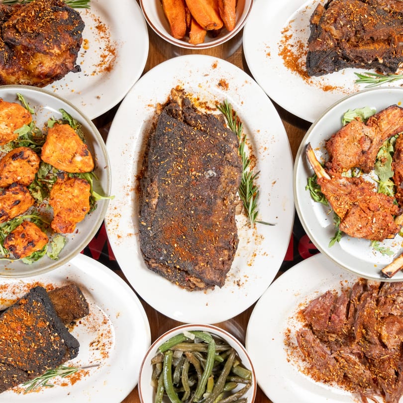 Overhead view of a feast of grilled meats, chicken, lamb chops, and vegetables on white plates.