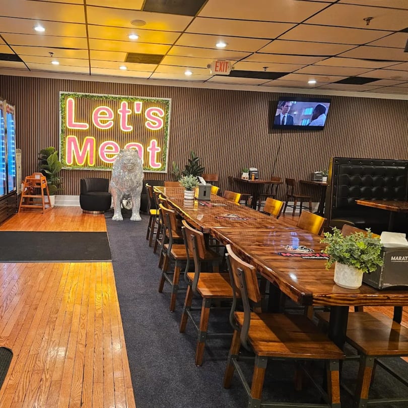 Interior of MeatVille Smoking restaurant with a long communal table, booths, and a "Let's Meat" neon sign.