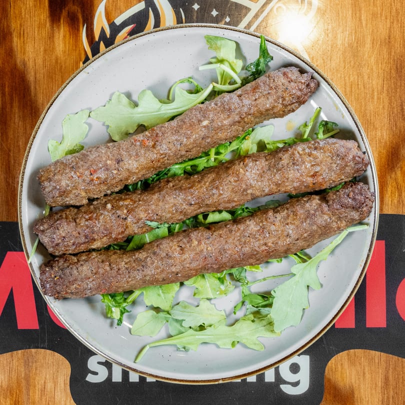 Three long, brown meat kebabs on a bed of green arugula, served on a light gray plate.