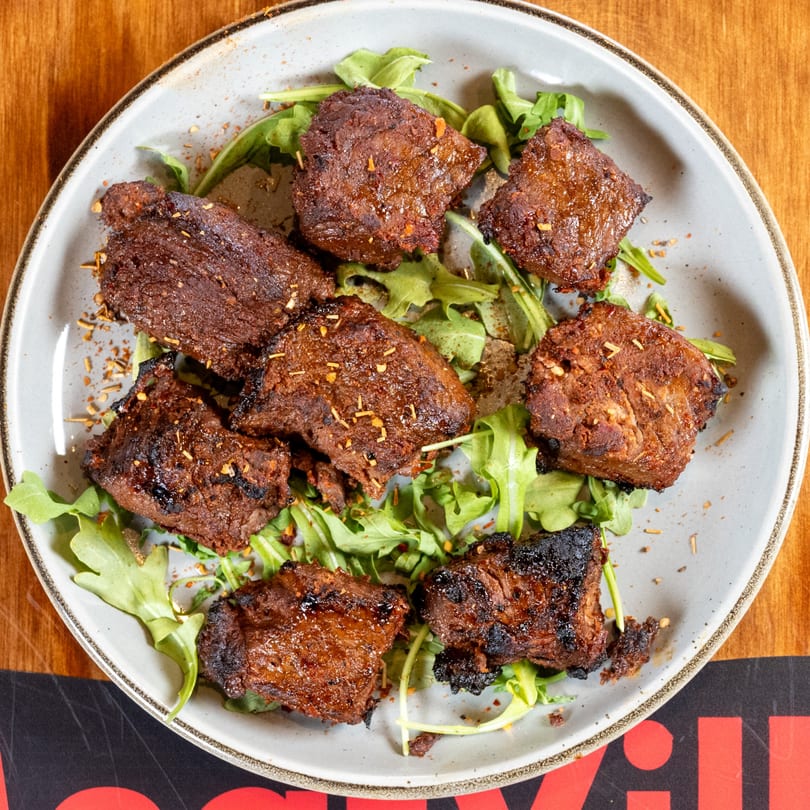 Top-down view of grilled meat pieces on a bed of arugula, sprinkled with spices on a gray plate.