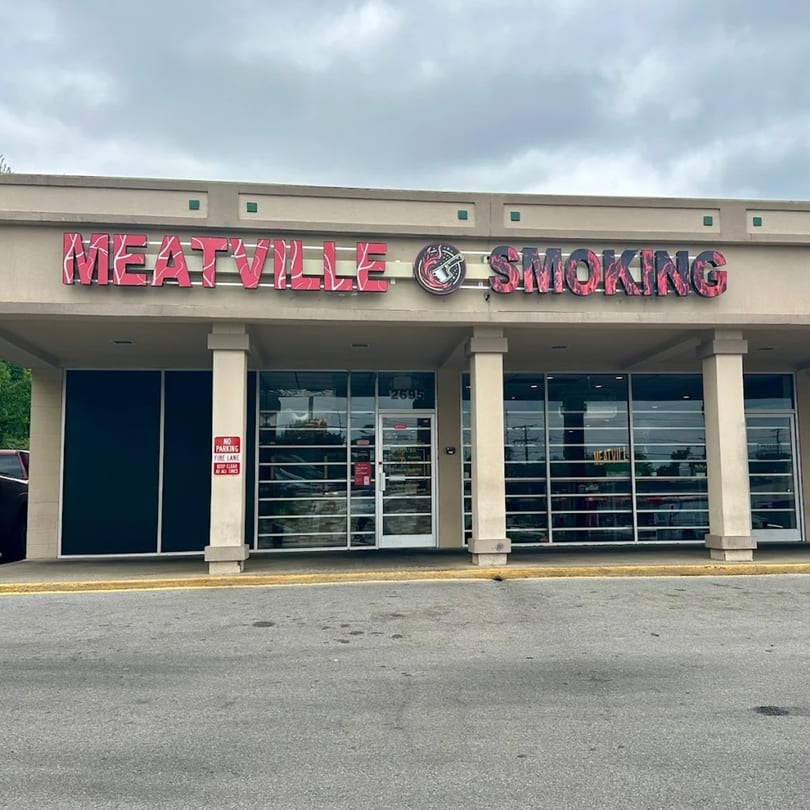 The exterior of MeatVille Smoking restaurant, featuring a tan facade, large windows, and a parking lot.
