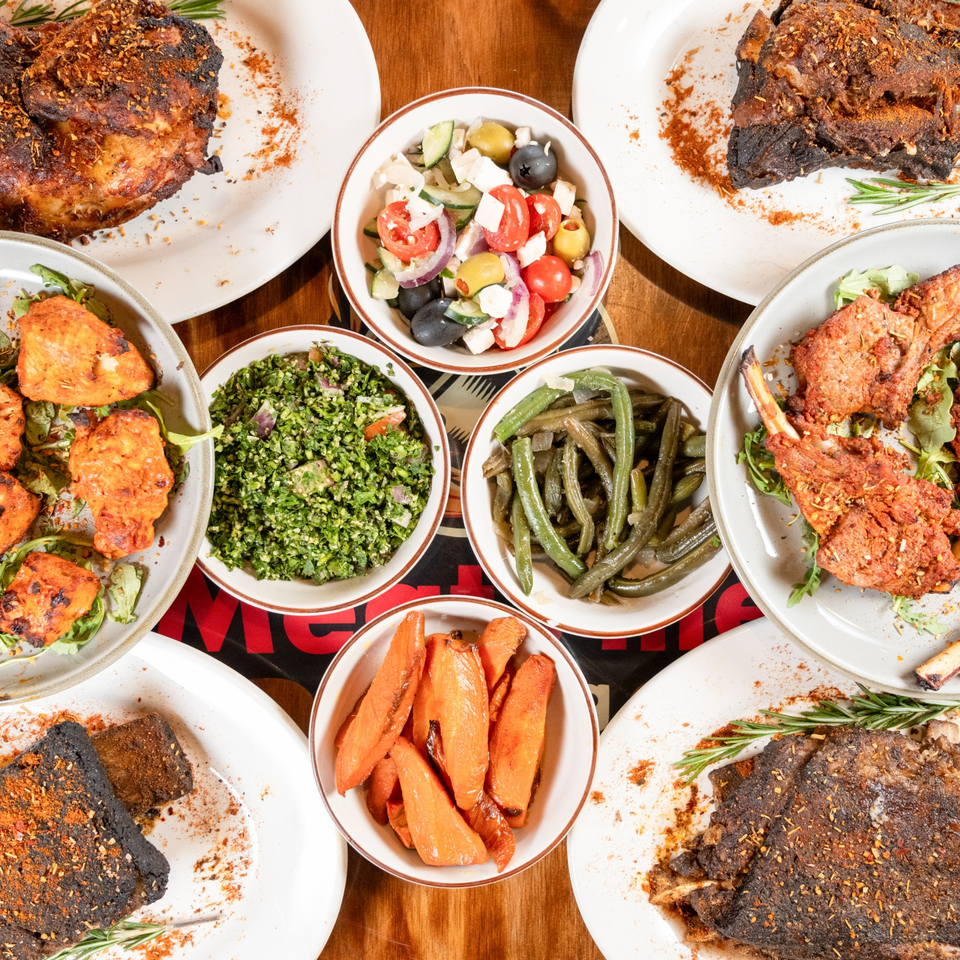 A feast of grilled meats, green beans, carrots, tabbouleh, and Greek salad on a wooden table.